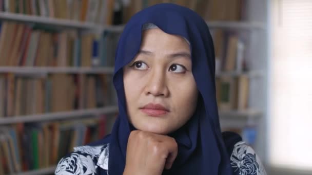 Asian muslim woman thinking and smiling while standing in front of bookshelf, education concept — Stock Video