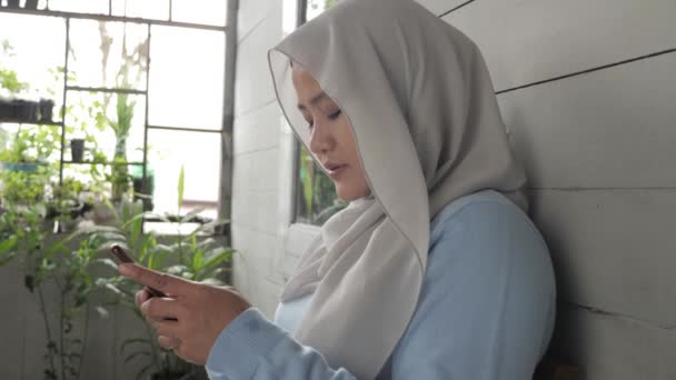 Asyalı Müslüman kadın evinin önünde oturuyor ve gülümsüyor. Mutlu hissediyorum. Kadın akıllı telefonuyla iletişim kuruyor. — Stok video
