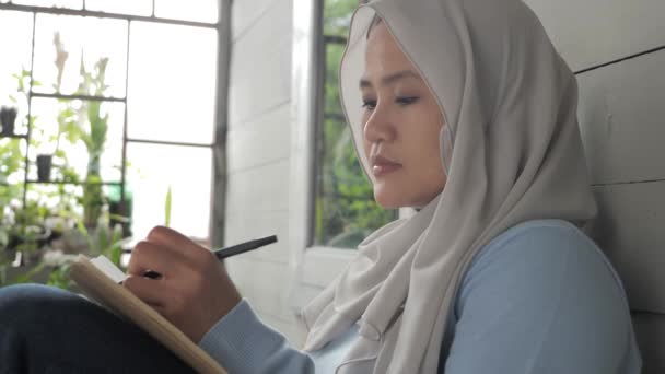 Schöne asiatische Muslimin sitzt auf ihrer heimischen Terrasse und schreibt an einem Buch — Stockvideo