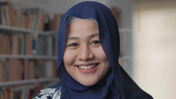 Счастливая азиатская мусульманка в хиджабе, улыбающаяся в камеру, студентка в библиотеке — стоковое видео