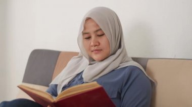 Güzel Asyalı Müslüman kadın kanepede otururken kitap okuyor, kız boş zamanlarında eğleniyor, neşeli bir ifade takınıyor.