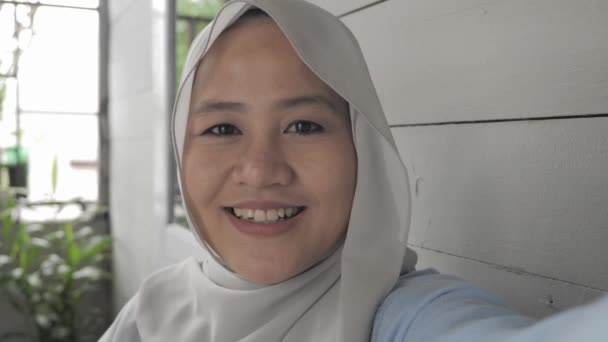 Schöne asiatische muslimische Dame beim Selfie-Porträt am Telefon oder beim Videoanruf, während sie vor ihrem Haus sitzt, fröhlich lächelnder fröhlicher Ausdruck — Stockvideo