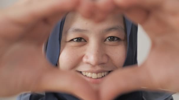 Portrait of happy cheerful Asian muslim woman smiled to camera while framing his face with heart shape made with her fingers — Stok Video