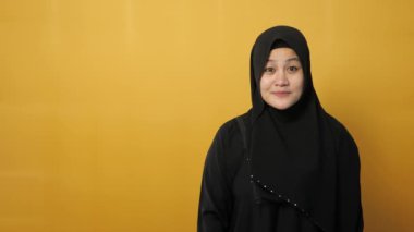 Mutlu Asyalı Müslüman kadın gülümsüyor ve kendi tarafındaki bir şeyi gösteriyor, sarı arkaplanda kopya alanı olan bir şeyi.