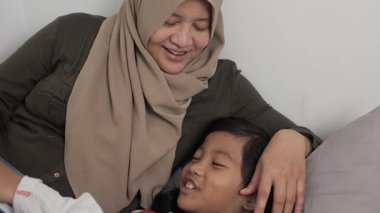 Asyalı Müslüman anne yatağında kızına masal okuyor, mutlu bir aile ilişkisi.