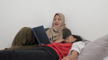 Asyalı Müslüman anne yatağında kızına masal okuyor, mutlu bir aile ilişkisi.