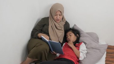 Asyalı Müslüman anne yatağında kızına masal okuyor, mutlu bir aile ilişkisi.