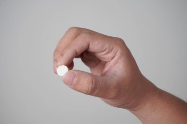 El, gri arka planda, parmaklarının arasında beyaz bir hap tutuyor.