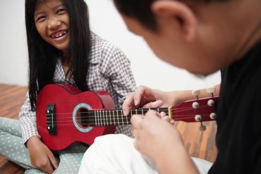 Asyalı bir baba küçük kızına evde gitar çalmayı öğretiyor, çocuğa müzik aletini tanıtıyor.