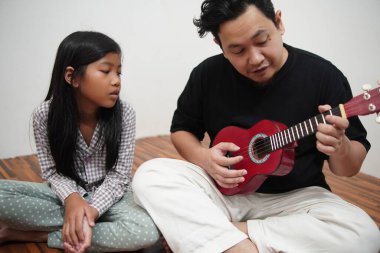 Asyalı bir baba küçük kızına evde gitar çalmayı öğretiyor, çocuğa müzik aletini tanıtıyor.