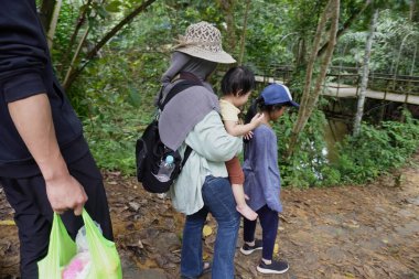 Asyalı Müslüman anne çocuklarıyla birlikte sabahın köründe yemyeşil bir parkta yürüyor. Mutlu bir aile açık hava aktivitesi yapıyor.