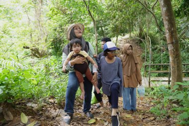 Asyalı Müslüman anne çocuklarıyla birlikte sabahın köründe yemyeşil bir parkta yürüyor. Mutlu bir aile açık hava aktivitesi yapıyor.