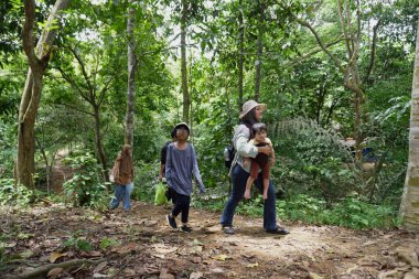 Asyalı Müslüman anne çocuklarıyla birlikte sabahın köründe yemyeşil bir parkta yürüyor. Mutlu bir aile açık hava aktivitesi yapıyor.