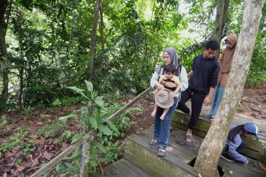 Asyalı Müslüman anne çocuklarıyla birlikte sabahın köründe yemyeşil bir parkta yürüyor. Mutlu bir aile açık hava aktivitesi yapıyor.