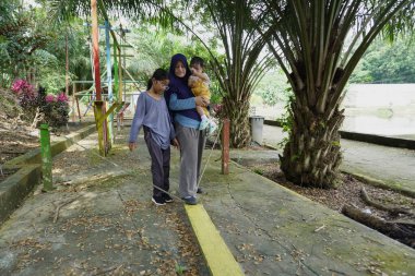 Asyalı Müslüman anne, güneşli bir sabahta küçük kızıyla parkta oynuyor. Mutlu bir aile, açık havada eğleniyor.