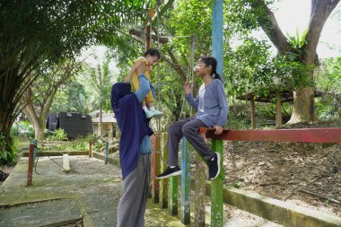 Asyalı Müslüman anne, güneşli bir sabahta küçük kızıyla parkta oynuyor. Mutlu bir aile, açık havada eğleniyor.