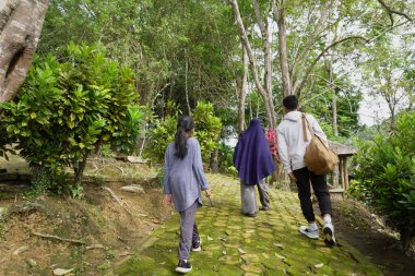 Asyalı Müslüman anne çocuklarıyla birlikte sabahın köründe yemyeşil bir parkta yürüyor. Mutlu bir aile açık hava aktivitesi yapıyor.