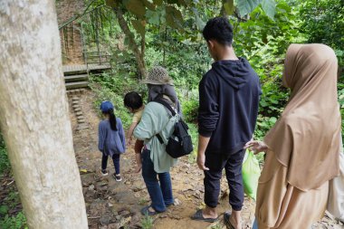 Asyalı Müslüman anne çocuklarıyla birlikte sabahın köründe yemyeşil bir parkta yürüyor. Mutlu bir aile açık hava aktivitesi yapıyor.