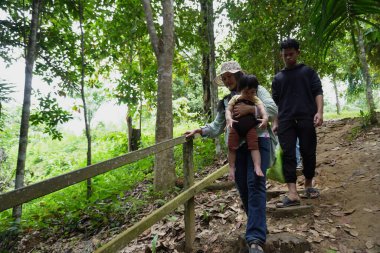 Asyalı Müslüman anne çocuklarıyla birlikte sabahın köründe yemyeşil bir parkta yürüyor. Mutlu bir aile açık hava aktivitesi yapıyor.