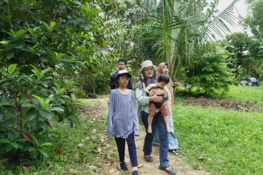 Asyalı Müslüman anne çocuklarıyla birlikte sabahın köründe yemyeşil bir parkta yürüyor. Mutlu bir aile açık hava aktivitesi yapıyor.