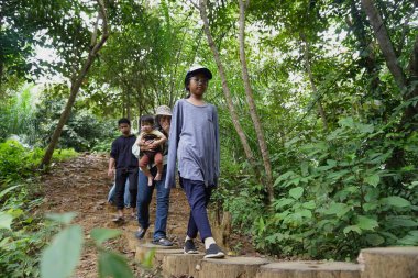 Asyalı Müslüman anne çocuklarıyla birlikte sabahın köründe yemyeşil bir parkta yürüyor. Mutlu bir aile açık hava aktivitesi yapıyor.