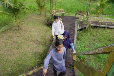 Zengin bir orman parkında ahşap bir köprüde yürüyen mutlu Asyalı anne ve çocuklar güneşli bir sabah yürüyüşü ve aile doğası gezintisinin keyfini çıkarıyorlar.