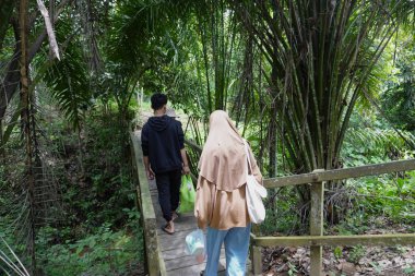 Mutlu Asyalı Müslüman aile ormanda ahşap köprüde yürüyor, anne ve çocuklar güneşli bir günde nehire ve şelaleye gidiyorlar, açık havada tatil yapıyorlar.