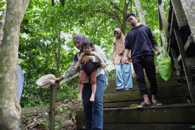 Zengin bir orman parkında ahşap bir köprüde yürüyen mutlu Asyalı anne ve çocuklar güneşli bir sabah yürüyüşü ve aile doğası gezintisinin keyfini çıkarıyorlar.