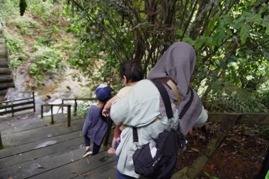 Mutlu Asyalı Müslüman aile ormanda ahşap köprüde yürüyor, anne ve çocuklar güneşli bir günde nehire ve şelaleye gidiyorlar, açık havada tatil yapıyorlar.