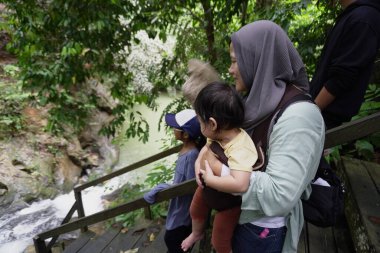 Mutlu Asyalı Müslüman aile ormanda ahşap köprüde yürüyor, anne ve çocuklar güneşli bir günde nehire ve şelaleye gidiyorlar, açık havada tatil yapıyorlar.