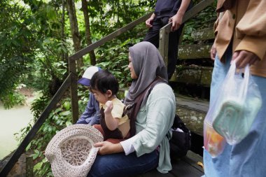Mutlu Asyalı Müslüman aile ormanda ahşap köprüde yürüyor, anne ve çocuklar güneşli bir günde nehire ve şelaleye gidiyorlar, açık havada tatil yapıyorlar.