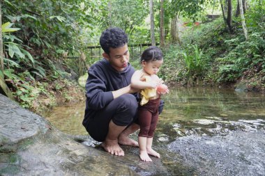 Asyalı kardeşler şelaleli küçük bir orman nehrinde oynuyor ve yüzüyorlar; mutlu çocuklar güneşli bir günde açık hava macerasından ve doğa keşiflerinden zevk alıyorlar.