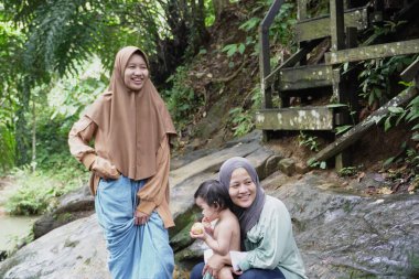 Mutlu Asyalı Müslüman anne tesettüre girip çocuklarıyla temiz bir orman nehrinde güneşli bir günde banyo yapıyor. Aile bağları doğada, açık havada yaz etkinliklerinde.