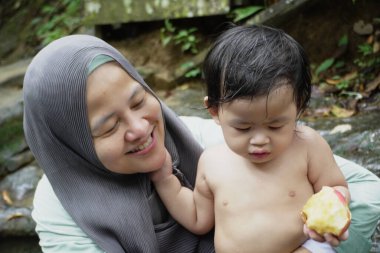 Mutlu Asyalı Müslüman anne tesettüre girip çocuklarıyla temiz bir orman nehrinde güneşli bir günde banyo yapıyor. Aile bağları doğada, açık havada yaz etkinliklerinde.
