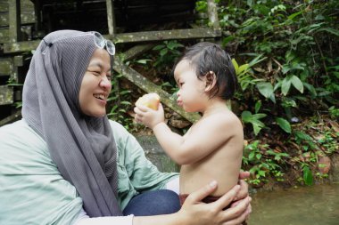 Mutlu Asyalı Müslüman anne tesettüre girip çocuklarıyla temiz bir orman nehrinde güneşli bir günde banyo yapıyor. Aile bağları doğada, açık havada yaz etkinliklerinde.
