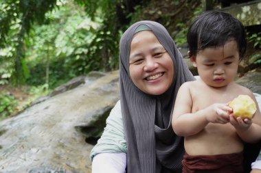 Mutlu Asyalı Müslüman anne tesettüre girip çocuklarıyla temiz bir orman nehrinde güneşli bir günde banyo yapıyor. Aile bağları doğada, açık havada yaz etkinliklerinde.