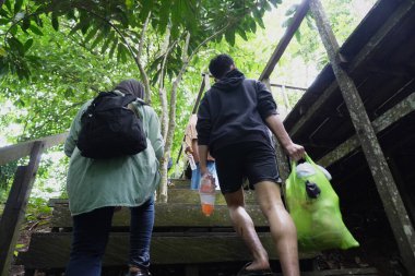 Mutlu Asyalı Müslüman aile ormanda ahşap köprüde yürüyor, anne ve çocuklar güneşli bir günde nehire ve şelaleye gidiyorlar, açık havada tatil yapıyorlar.