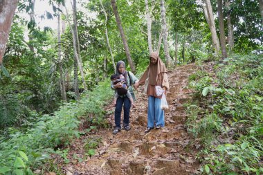 Asyalı Müslüman anne genç kızıyla el ele tutuşuyor ve bebeği taşıyıcıyla ormanda yürürken taşıyor, güneşli bir sabah ailece doğa yürüyüşü yapıyor.