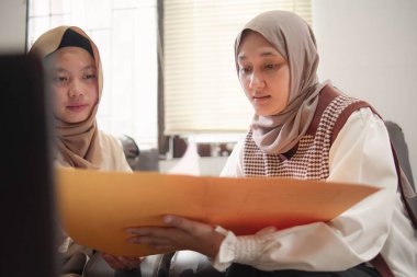 İki güzel Asyalı genç Müslüman iş kadını modern bir ofiste çalışırken gülümseyen bir klasörde belgeleri tartışıyor.