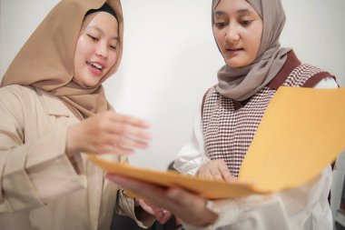 İki güzel Asyalı genç Müslüman iş kadını modern bir ofiste çalışırken gülümseyen bir klasörde belgeleri tartışıyor.