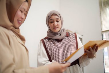 İki güzel Asyalı genç Müslüman iş kadını modern bir ofiste çalışırken gülümseyen bir klasörde belgeleri tartışıyor.