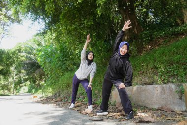 Yeşil bir parkta güneşli bir günde, tesettür takan ve ısınan iki genç Müslüman kadın. Sağlıklı yaşam tarzı, arkadaşlık ve doğal ışık ile uygunluk kavramı