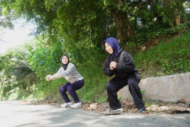 Yeşil bir parkta, tesettürlü, zıplama egzersizi yapan, güneşli bir günde gülümseyen ve gülen iki genç Müslüman kadın. Aktif yaşam tarzı, arkadaşlık ve doğal ışık ile uygunluk kavramı