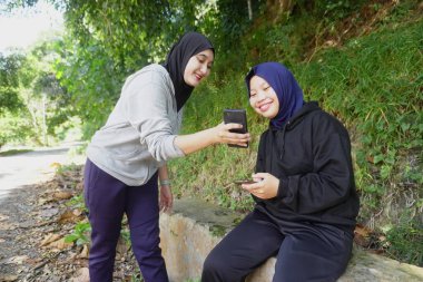 Açık havada egzersiz yaptıktan sonra tesettür takan iki genç Müslüman kadın güneşli bir günde yeşil bir parkta mutlu bir şekilde gülüyor ve akıllı telefonu paylaşıyor. Arkadaşlık, teknoloji ve doğal yaşam tarzı kavramı.