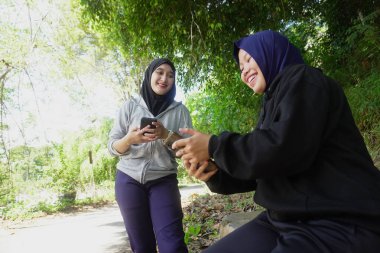 Açık havada egzersiz yaptıktan sonra tesettür takan iki genç Müslüman kadın güneşli bir günde yeşil bir parkta mutlu bir şekilde gülüyor ve akıllı telefonu paylaşıyor. Arkadaşlık, teknoloji ve doğal yaşam tarzı kavramı.