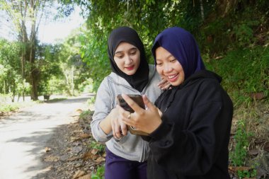Açık havada egzersiz yaptıktan sonra tesettür takan iki genç Müslüman kadın güneşli bir günde yeşil bir parkta mutlu bir şekilde gülüyor ve akıllı telefonu paylaşıyor. Arkadaşlık, teknoloji ve doğal yaşam tarzı kavramı.