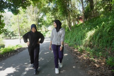 Güneşli bir günde ağaçlarla dolu yeşil bir park yolunda sabah egzersizi için birlikte yürüyen tesettür giyen iki genç Müslüman kadın. Aktif yaşam tarzı, arkadaşlık ve doğal ışık ile sağlıklı yaşam konsepti