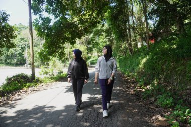 Güneşli bir günde ağaçlarla dolu yeşil bir park yolunda sabah egzersizi için birlikte yürüyen tesettür giyen iki genç Müslüman kadın. Aktif yaşam tarzı, arkadaşlık ve doğal ışık ile sağlıklı yaşam konsepti