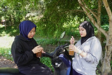 İki genç Müslüman kadın tesettür takıyor ve güneşli bir günde soğuk meyve suyu içip gülümserken gölgeli yeşil ağaçların altında motosikletin altında dinleniyorlar. Hayat tarzı, arkadaşlık ve sağlıklı yaşam kavramı, doğal ışık ile tazeleniyor.