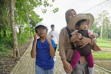 Asyalı Müslüman anne, çocuklarıyla yemyeşil bir parkta ağaçlık bir patika boyunca yürürken bebeğini taşıyor. Aile, sağlıklı bir yaşam tarzını, ebeveynliği, birlikteliği ve her şeyi temsil eden doğal bir çevrede huzurlu bir doğa yürüyüşü yapmayı seviyor.